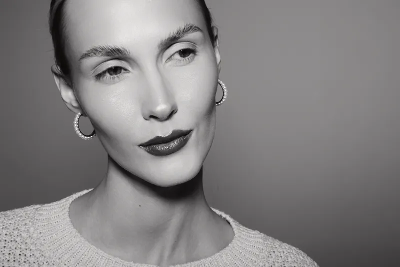 Close-up portrait of a slender Caucasian woman with light skin and short hair wearing earrings and a knitted sweater, looking sideways in a monochrome studio setting.