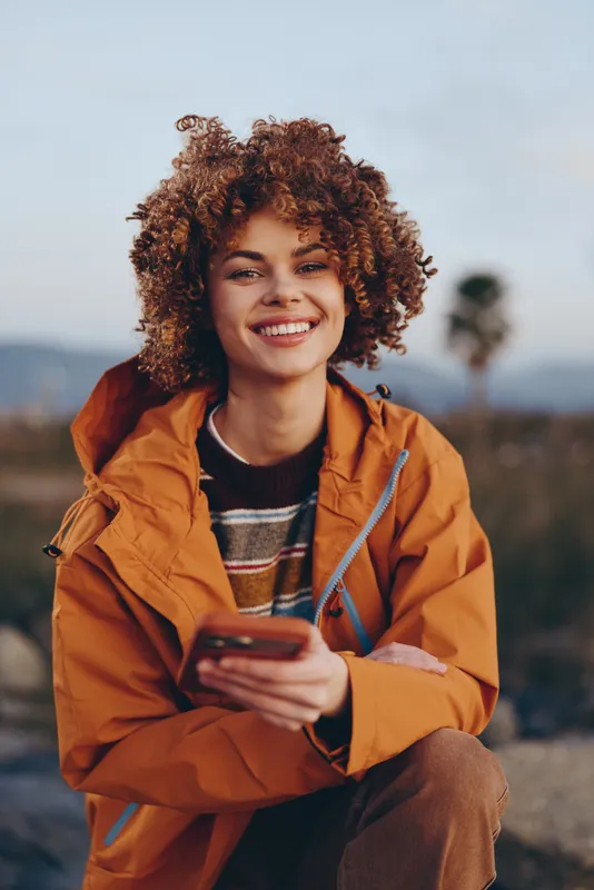 Woman lifestyle outdoors, holding smartphone, curly hair and smiling portrait, orange jacket against mountains and landscape, travel vibe, natural daylight, shallow depth of field