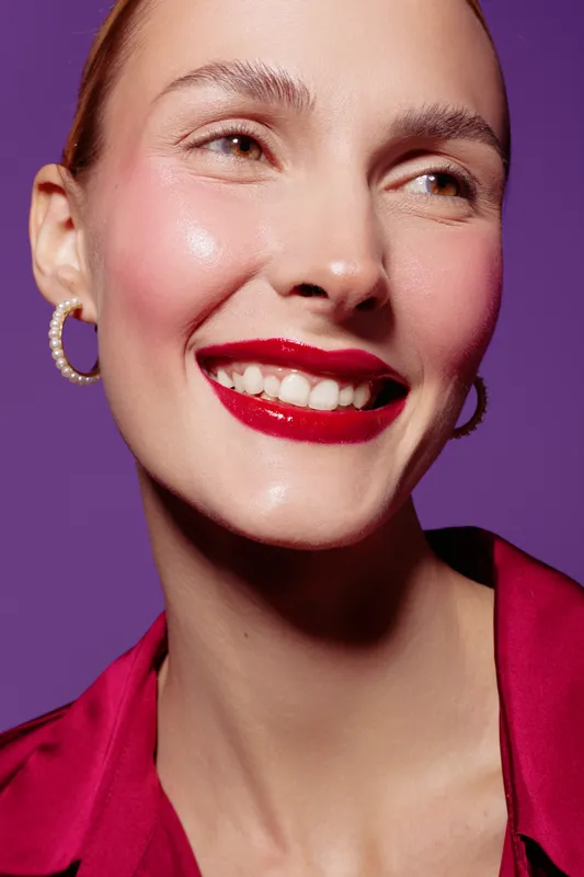 Smiling woman with red lipstick and hoop earrings wears a red blouse against purple background. Close-up portrait with natural skin glow and joyful expression. Beauty and fashion concept.