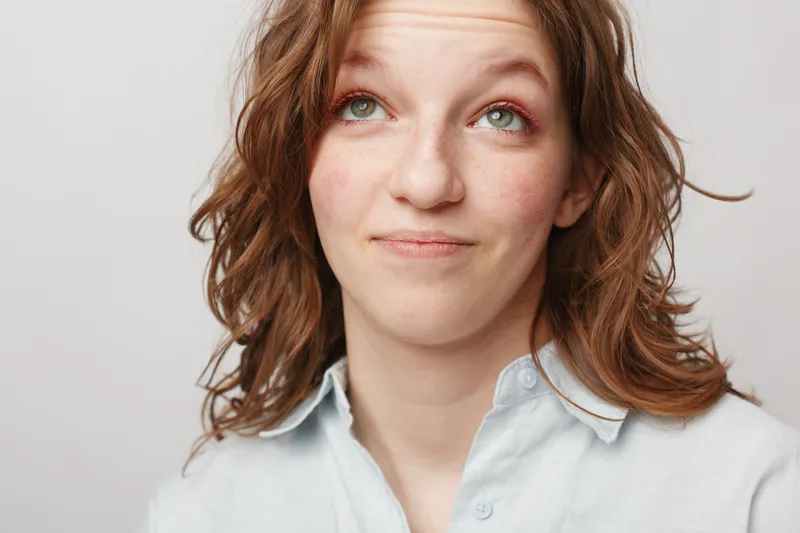 Young woman with curly hair looks up with a thoughtful expression wearing a light blue shirt. Studio portrait on white background. Concept of contemplation and casual style.
