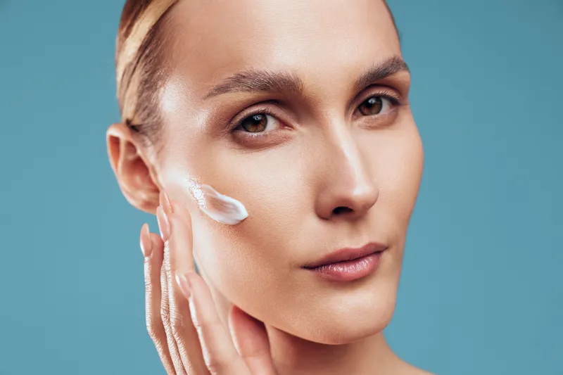 Young woman applies moisturizing cream on her cheek skin with fingers close up portrait isolated on blue background with natural makeup and healthy facial expression.