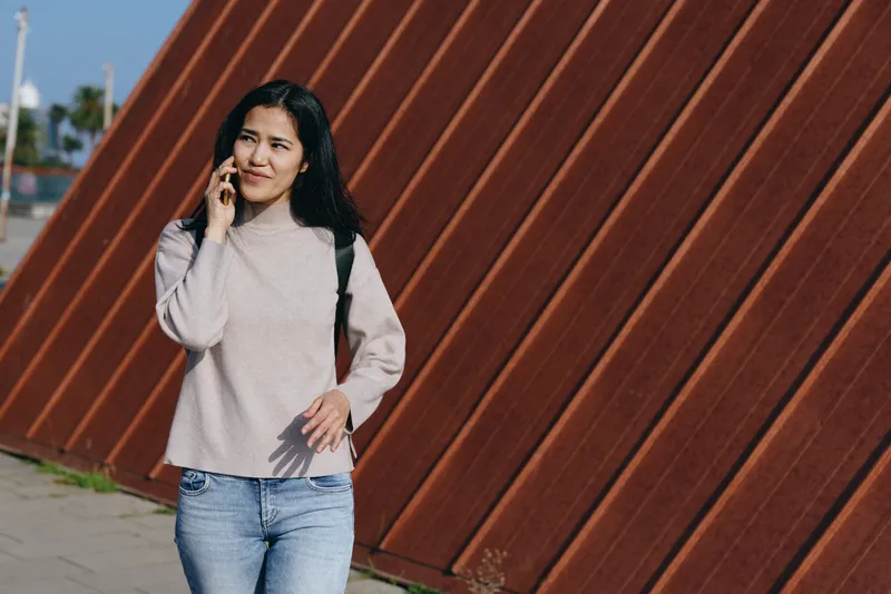 Woman on phone while walking along a rusted wall, casual sweater and jeans, outdoor street scene capturing fashion and mobility, urban lifestyle commentary and everyday communication