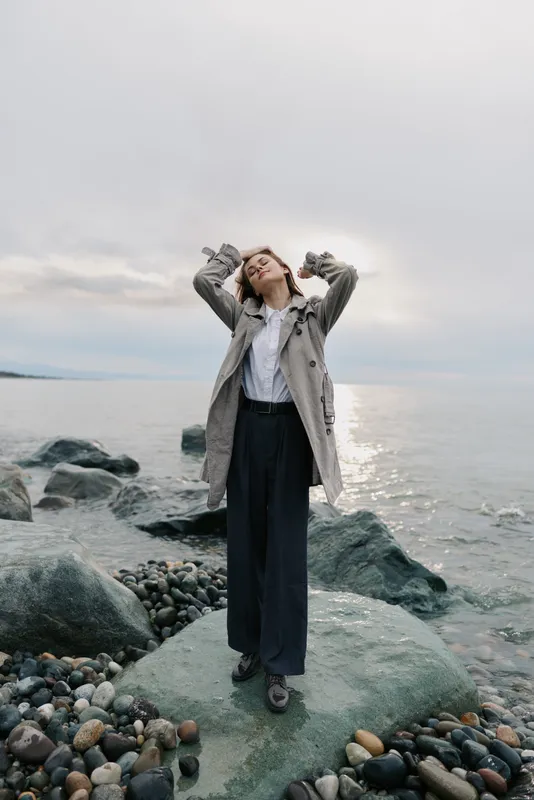 Young woman stands on rocky shore wearing coat and pants with eyes closed and hands raised. Soft natural light, cloudy sky, and calm water create peaceful mood on beach.