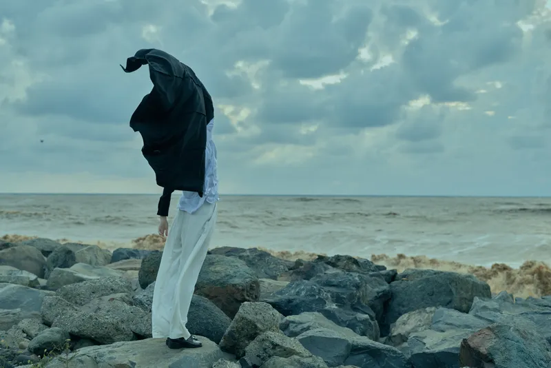 Figure on rocky shoreline with windblown jacket hood, sea and cloudy horizon, conveying solitude, exploration, and resilience in a dramatic coastal landscape