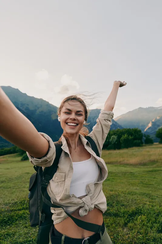 A young woman with a backpack raises her arms in a sunny mountain meadow, smiling as she enjoys an adventurous hike and fresh air, feeling free in the moment.