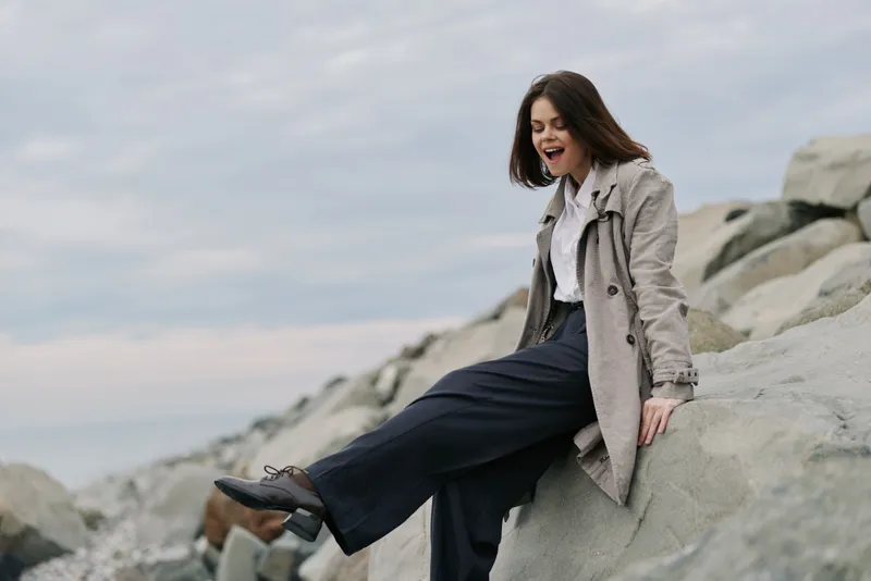 Young woman in coat and trousers sits on rocks near the sea on cloudy day. She looks joyful and relaxed, enjoying the natural outdoor environment by the coast with casual style.