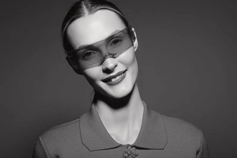 Portrait of a smiling Caucasian woman with pale skin wearing futuristic sunglasses and a textured sweater on a dark background.