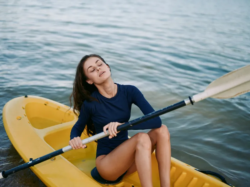 A relaxed woman sits in a yellow kayak on calm water, eyes closed, enjoying the breeze as she rests with a paddle across her lap, capturing a serene outdoor moment.