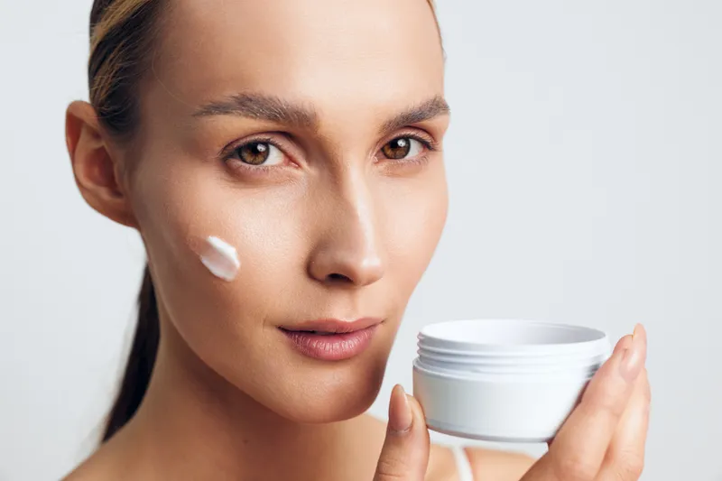 Young woman applying face cream on cheek with smooth skin, holding white jar, skincare routine, natural makeup, clean background, focused expression, beauty concept