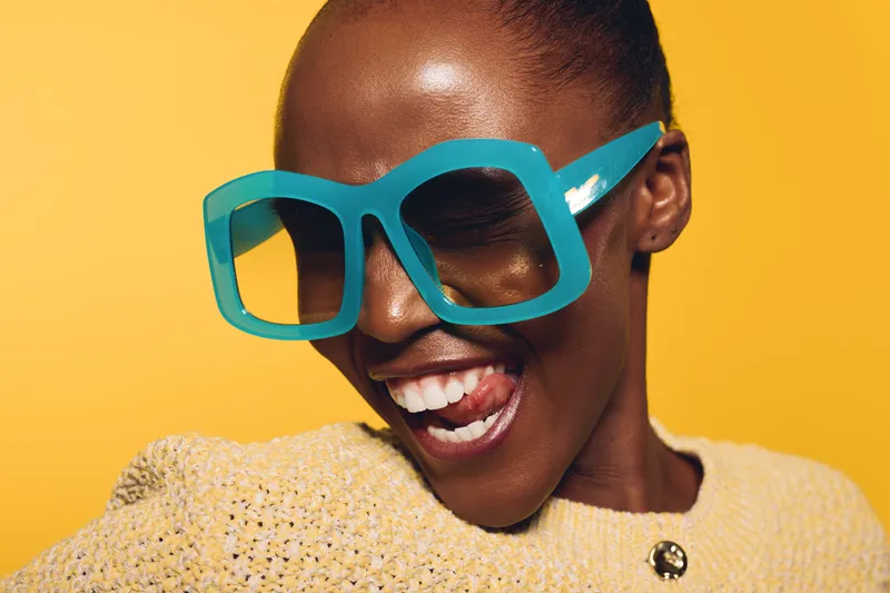 Close-up of a happy dark skinned African woman wearing oversized turquoise sunglasses and a textured yellow sweater. She smiles with tongue out on bright yellow background. Fun fashion concept.
