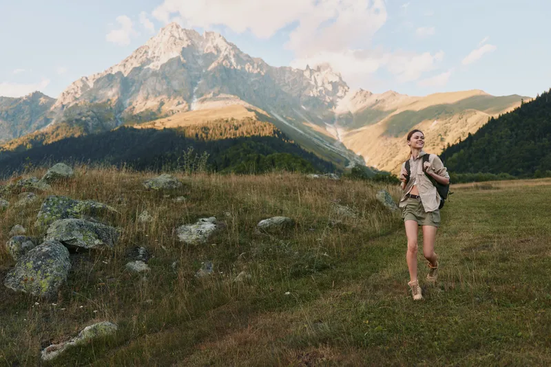 A young woman hiker in shorts and jacket runs across a wide alpine meadow with rugged mountains in the background, capturing an energetic moment of outdoor exploration.
