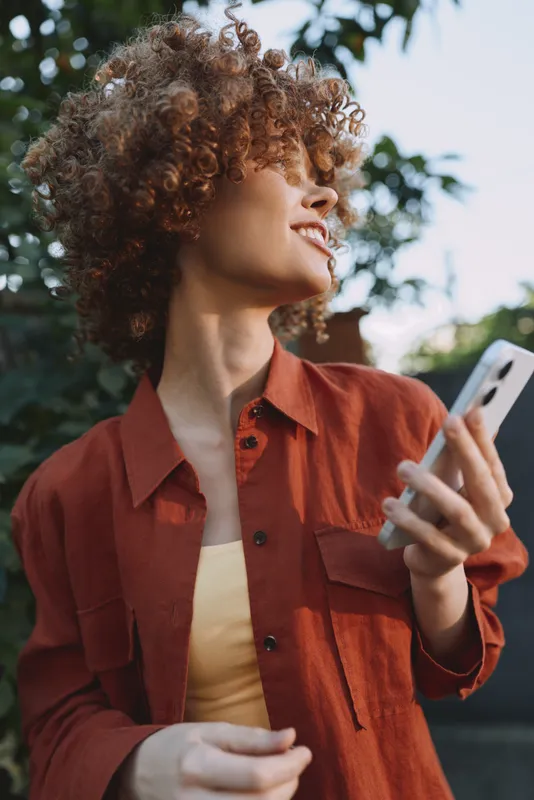 Young woman with curly hair checks her phone outdoors, smiling softly as warm sunlight filters through leafy trees, capturing a casual lifestyle moment