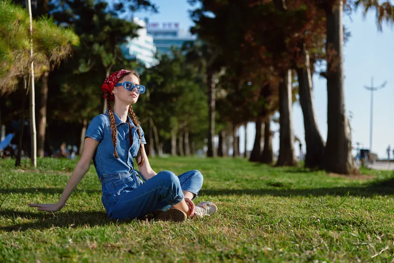 Lifestyle woman in modern boho-western denim street style sits on grass outdoors, enjoying sunny weather with casual vintage film color tones and relaxed summer vibe