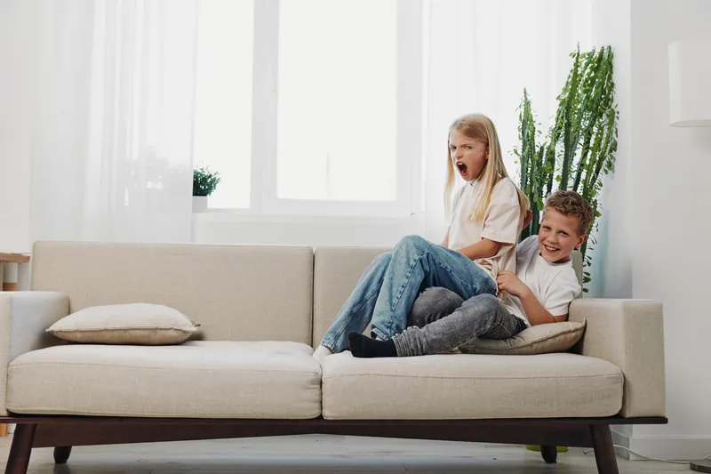 Playful children laughing and having fun on a modern beige sofa in a bright living room with natural light and green plants. Concept of childhood joy, family bonding, and home comfort.