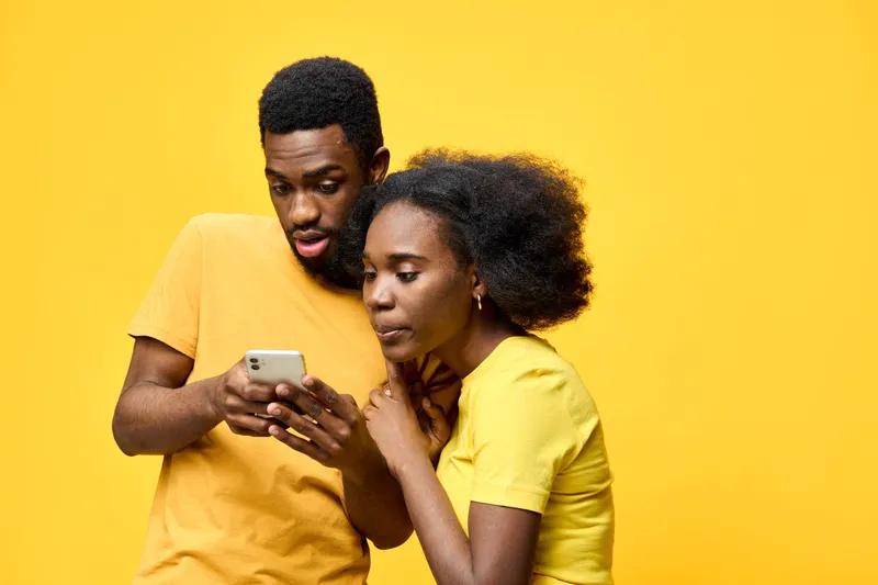 A joyful couple of African descent in matching yellow shirts, engaged with a smartphone against a bright yellow background, conveying intimacy and connection