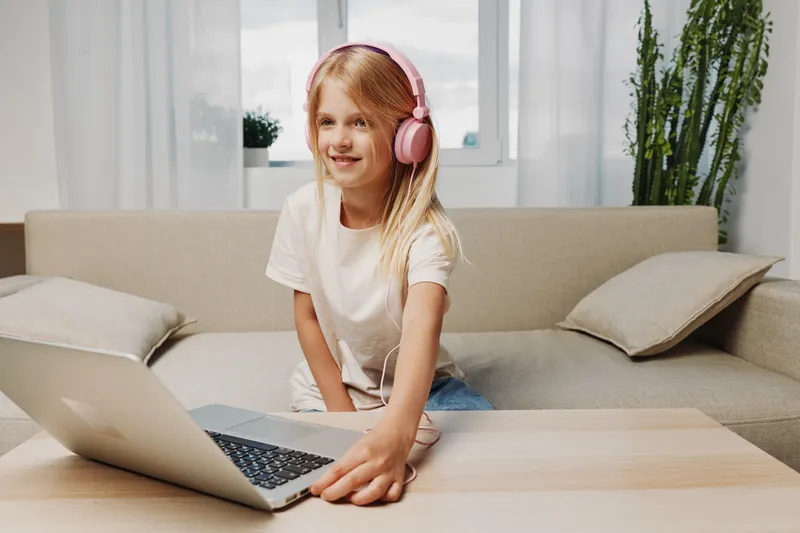 Smiling girl with pink headphones uses laptop on wooden table in bright living room. Concept of online learning, technology, childhood and digital education at home.