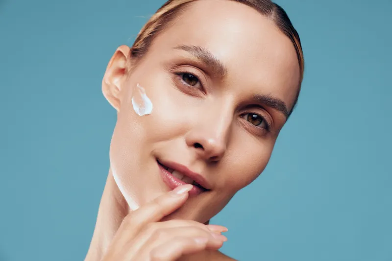 One person, a young woman with brunette hair, applies facial cream on her cheek in a studio with a blue background, capturing a calm skincare moment in a close up portrait.