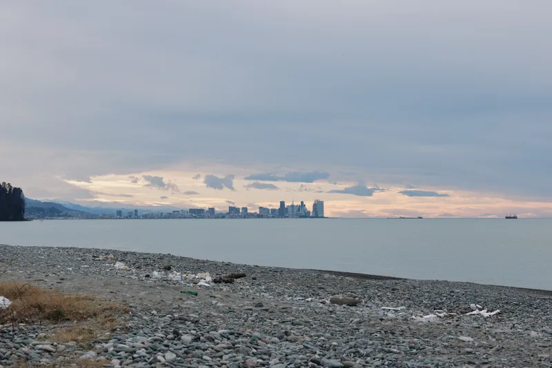 Beach and shoreline meet calm water with distant cityscape on the horizon under moody clouds at sunset, creating a tranquil coastal scene with muted tones and gentle light across the shore.
