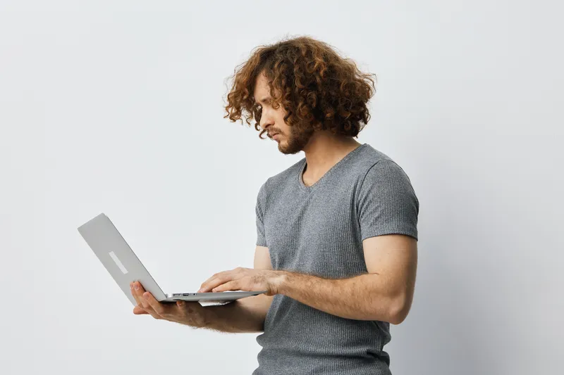 A young man with curly hair in a gray shirt using a laptop, showcasing concentration and focus in a minimalistic setting Perfect for business or lifestyle themes