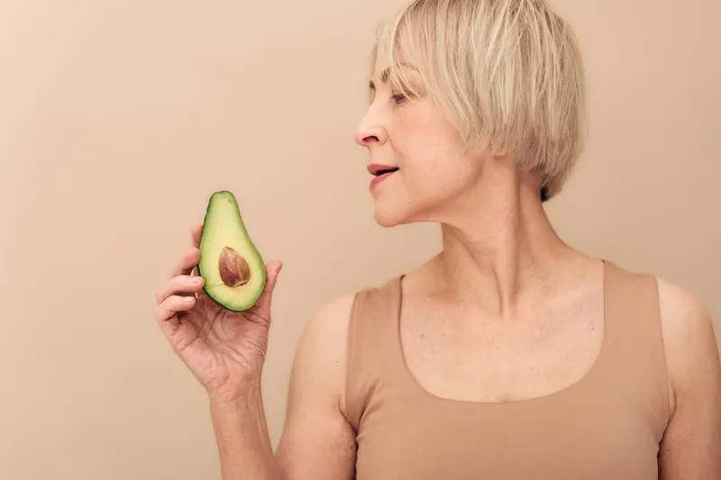 Mature woman with short blonde hair holds half of ripe avocado near face on beige background. Concept of healthy eating, natural food, nutrition, and wellness for middle aged people.