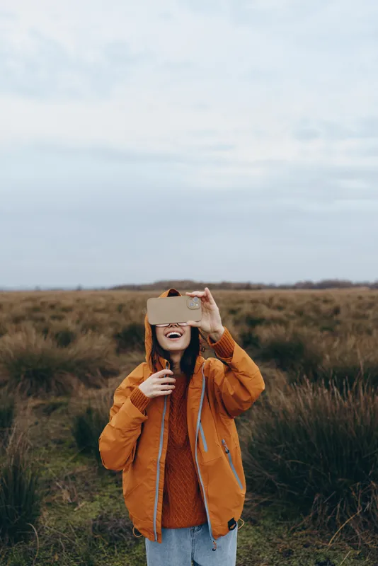 Woman lifestyle outdoors in nature field, smiling and holding a smartphone like a viewer, wearing an orange jacket, autumn grass landscape, candid fashion mood, soft daylight