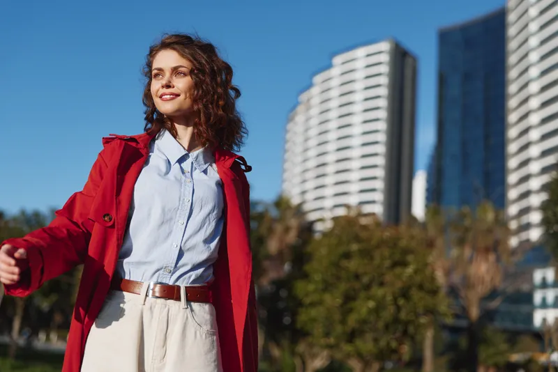 Young woman with curly hair wearing a red coat and white pants enjoys a sunny day in the city with modern buildings in the background.