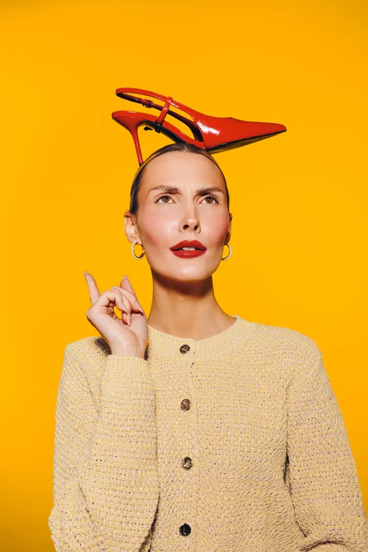 Thoughtful woman in beige sweater balances red high heel shoe on her head against bright yellow background. Concept for fashion, creativity, and unique style expression.