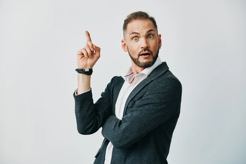 An assertive business professional stands against a solid colored backdrop, capturing a surprised, alert moment as he points upward with one finger.