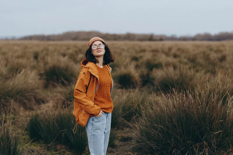Woman lifestyle outdoors field grass autumn hat glasses jacket, orange jacket and knit hat, denim jeans, standing in tall grass, calm smile, looking at camera, countryside landscape, autumn breeze,