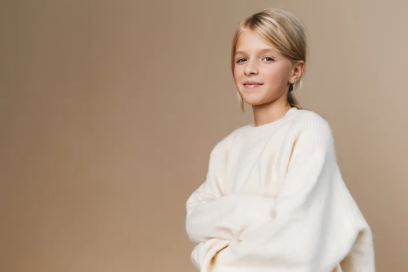Portrait of a young girl in a soft ivory sweater, standing beside a warm beige backdrop with a calm, confident smile and relaxed posture, enhanced by subtle lighting and gentle energy.