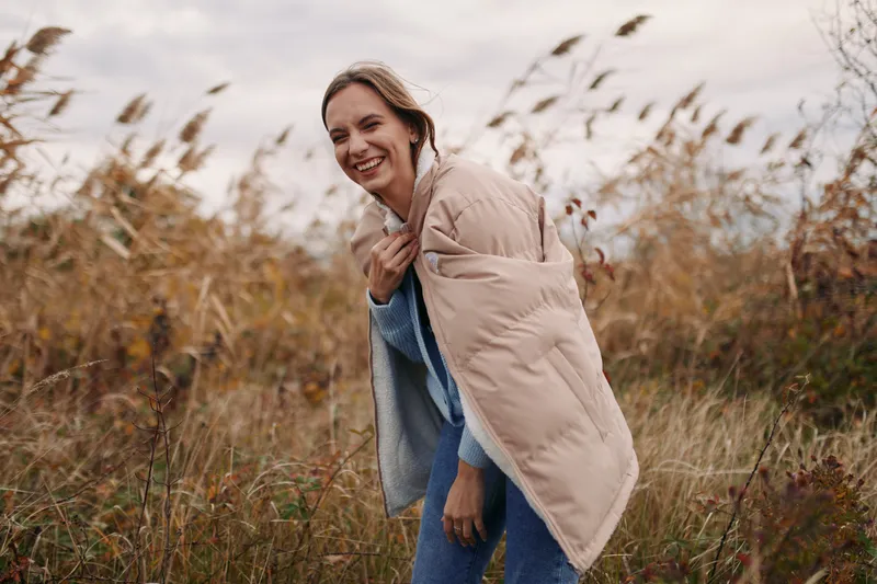 A candid portrait of a smiling woman wrapped in a soft beige coat, standing in tall grasses, conveying warmth, authenticity, and everyday charm in natural light.