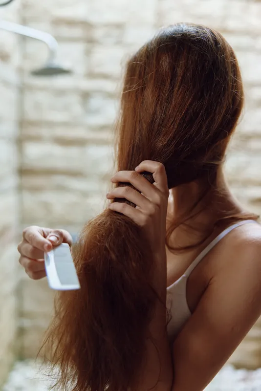 Woman combing long brown hair indoors with natural light, wearing white tank top, focusing on hair care and grooming routine, calm and intimate atmosphere.
