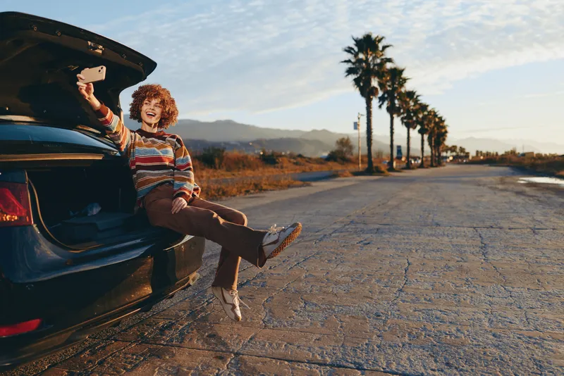 woman lifestyle outdoors, sitting on open car trunk beside a quiet roadside at sunset, casual striped shirt and denim jeans, relaxed smile, warm light, palm trees, distant wide mountains