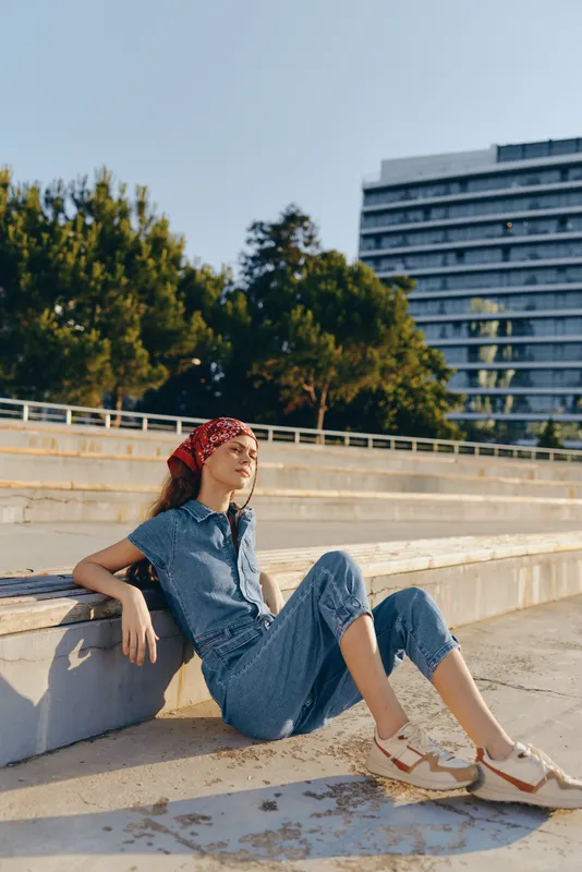relaxed woman wearing denim jumpsuit and red bandana sitting outdoors on concrete steps in sunny urban park with trees and building background