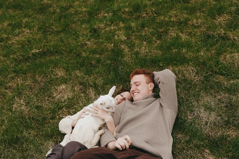 Goat, kid, man outdoors, relaxing moment as they lie on grass, cozy sweater, gentle smile, sunlight, tender interaction, nature backdrop