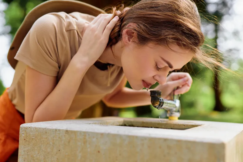 Woman drinking from public fountain in park, leaning over stone basin to sip water. Outdoor summer scene with backpack strap, casual clothing, wristwatch, natural greenery and sunlight.