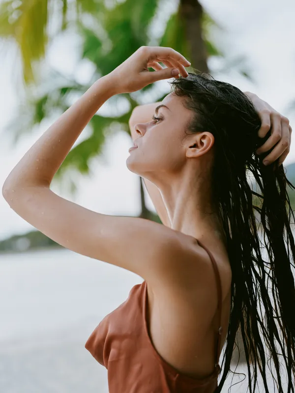 Relaxed woman with wet hair enjoying tropical outdoors, wearing a sleeveless top, with green palm background and natural sunlight.