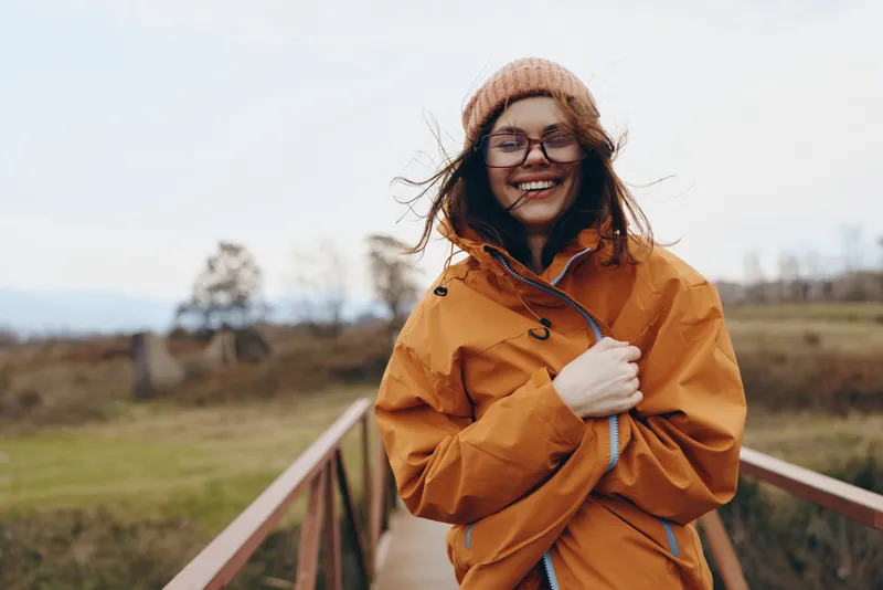 Woman lifestyle outdoors smiling orange raincoat glasses bridge countryside autumn daylight, holding coat collar, standing by railing, rural landscape, travel leisure mood