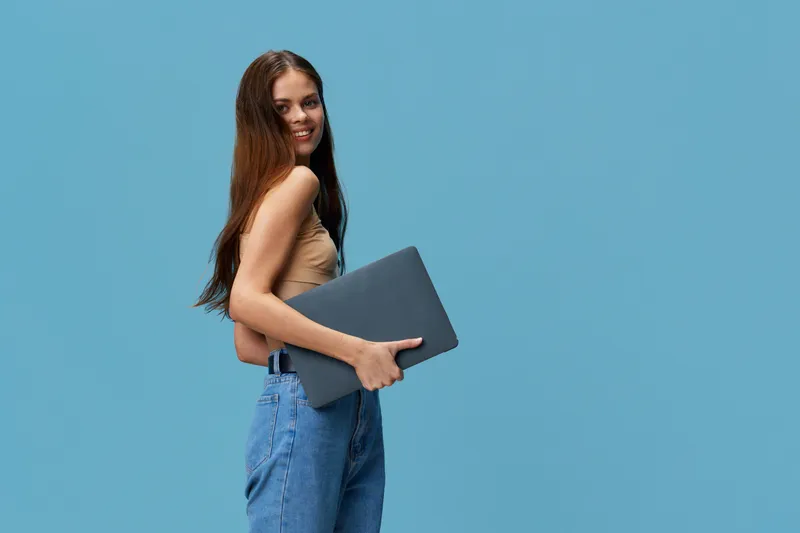 smiling woman holding laptop wearing casual beige top and blue jeans against plain blue background with long brown hair and relaxed posture