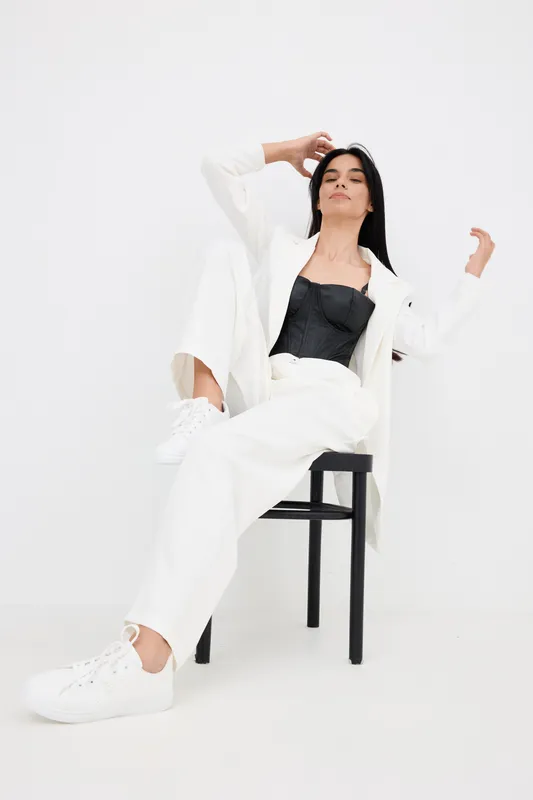 relaxed woman in white suit sitting on black chair wearing sneakers and black top, stylish outfit and casual pose on plain white background