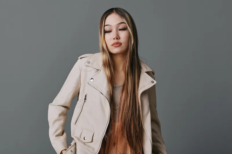 Young woman with long braided hair wearing a beige jacket standing against gray studio background. Fashion portrait with casual style and closed eyes for lifestyle or beauty concept.