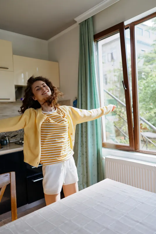Young woman with curly hair stretches her arms in a bright kitchen beside a large window, wearing a yellow cardigan over a striped shirt and white shorts, conveying a fresh, home lifestyle moment