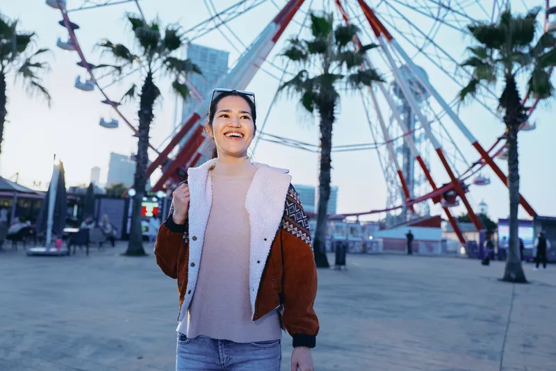 Woman smiling in a cozy jacket stands outdoors near a ferris wheel and palm trees, city vibe at dusk, casual fashion and travel mood, candid street photography scene
