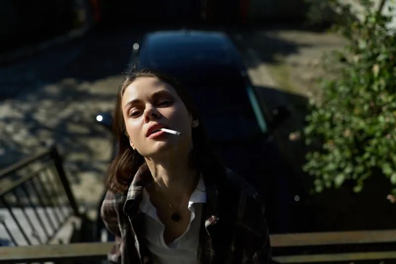 Young woman smoking outdoors, casual portrait on a shaded street with moody natural lighting, portraying everyday lifestyle and relaxed urban attitude
