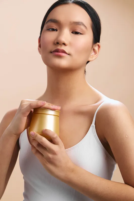 asian woman in a white tank top holds a golden skincare jar, posed in soft studio light. she smiles gently, presenting a calm, confident, minimal beauty moment.