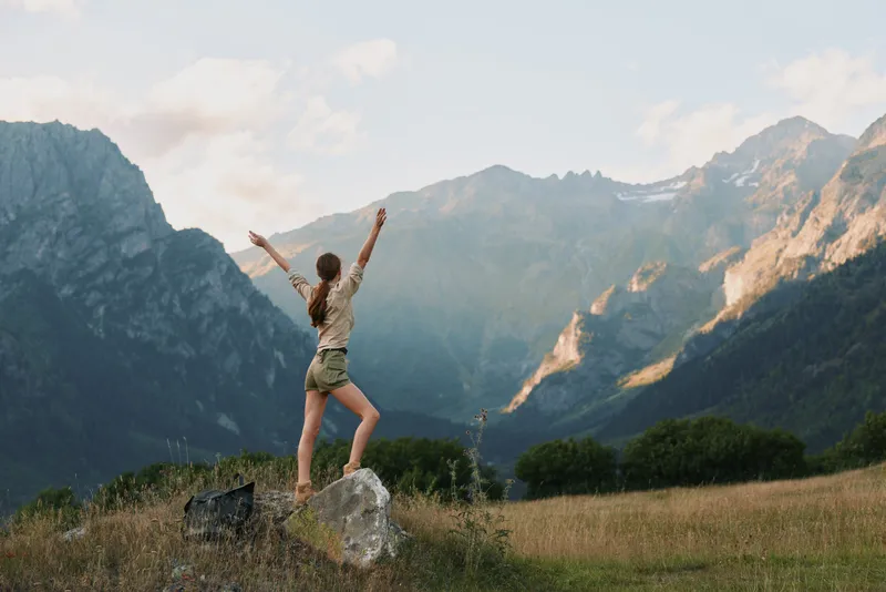 Cheerful woman outdoors, standing on a rock with arms raised, mountain valley backdrop, sunny day, carefree mood, nature inspired lifestyle
