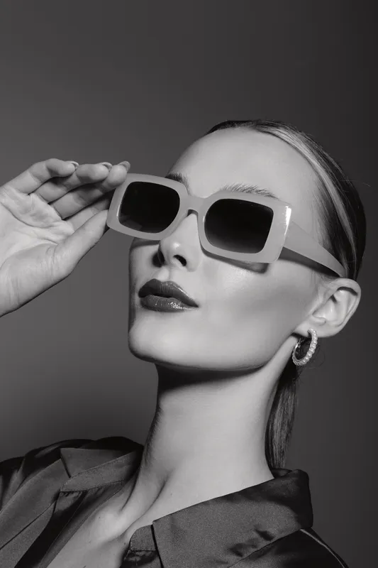 Stylish Caucasian woman with fair skin wears large sunglasses and hoop earrings, posing confidently in black and white studio portrait. Fashion and elegance concept.