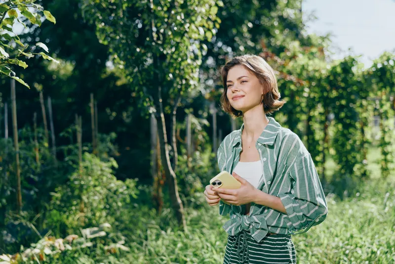 Woman outdoors portrait with a cheerful smile in a sunlit garden, casual attire, holding a phone, she radiates relaxed confidence and natural charm, inviting calm and friendly vibes.