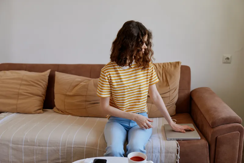 Woman seated on a sofa at home, relaxed with striped shirt and jeans, glancing at a tablet beside coffee, cozy living room scene.