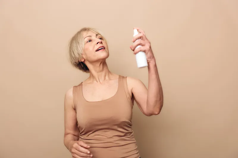 Mature woman in beige tank top sprays facial mist on her face, enjoying skincare routine. Neutral background emphasizes self-care, hydration, and natural beauty for older adults.